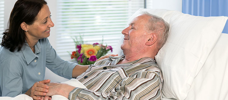 Female palliative care provider smiles while gently holding an elderly man's hand while he is lying comfortably in bed.
