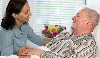 Female palliative care provider smiles while gently holding an elderly man's hand while he is lying comfortably in bed.