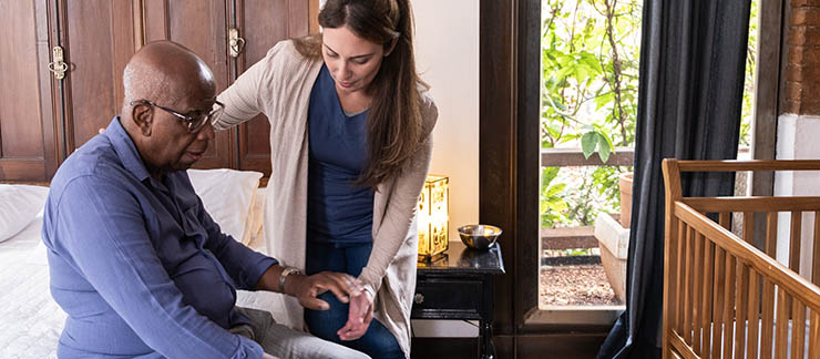 A female palliative caregiver helping a senior man standing up from bed at home.