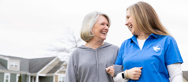 A Visiting Angels female caregiver walks arm and arm smiling with a senior woman outside of her home.