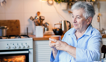 Senior woman sips coffee in kitchen while food is cooking in the oven.