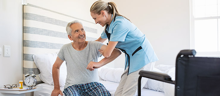Smiling female home care worker assisting senior man to get up from bed and move towards wheelchair at home.