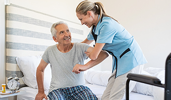 Smiling female home care worker assisting senior man to get up from bed and move towards wheelchair at home.