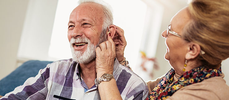 Woman helps her senior husband attach his hearing aid to his ear at home.