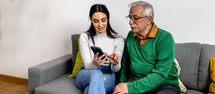 Mid-aged female sitting on couch with elderly man at home showing him how to safely answer incoming calls on his cell phone.