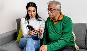 Mid-aged female sitting on couch with elderly man at home showing him how to safely answer incoming calls on his cell phone.