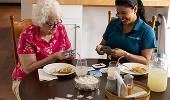 Elderly woman with cell phone sits at breakfast table with a female caregiver who is smiling looking at her phone.