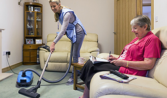 Senior woman sits on sofa in her home while female home care worker vacuums the carpet.