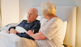 Disoriented senior man is comforted in bed by his wife who compassionately holds his arm.