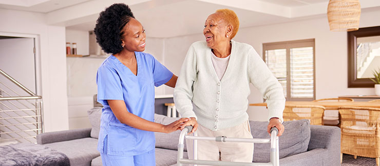 Female caregiver helps senior woman with a cane to walk around her home.