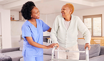 Female caregiver helps senior woman with a cane to walk around her home.