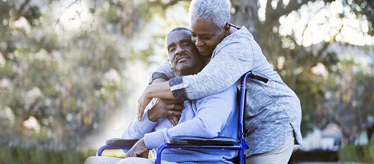 Senior woman hugs husband in wheelchair outside.