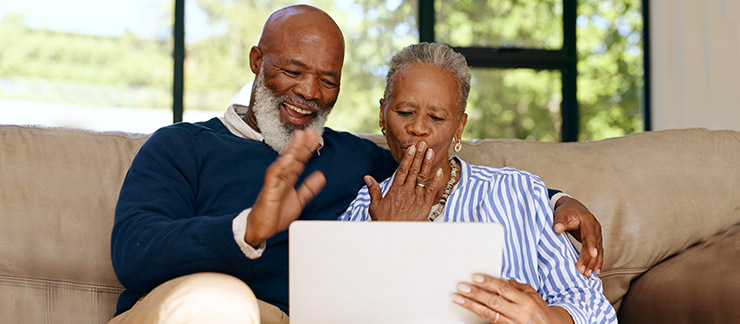 Senior couple sitting on a sofa with a computer tablet waving to family members who are away on vacation.