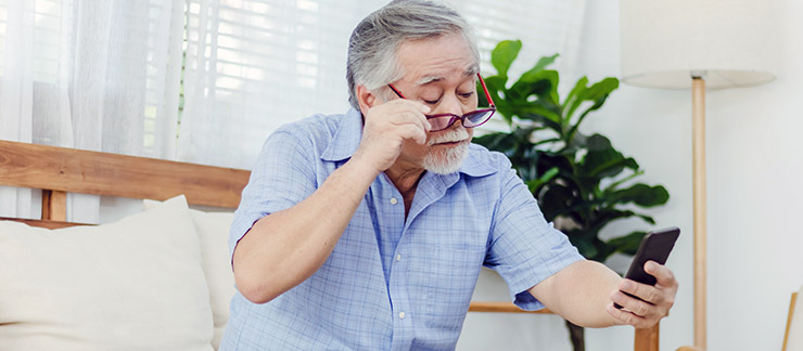 Senior male with glasses is having a hard time reading the screen of his cell phone while sitting on couch at home.