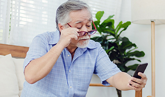 Senior male with glasses is having a hard time reading the screen of his cell phone while sitting on couch at home.
