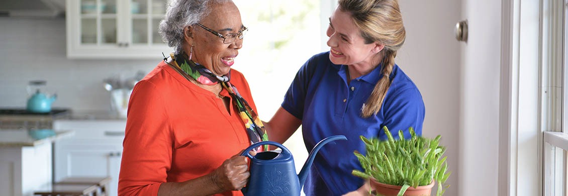 Mid-aged woman takes provide in her caregiver job by helping an elderly woman at home water a plant.