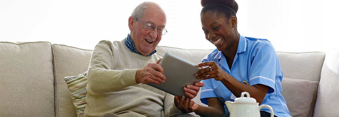 Elderly man learns to use a computer tablet from a helpful home care worker.