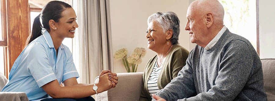 Female caregiver chats with a senior couple at home sitting on sofa