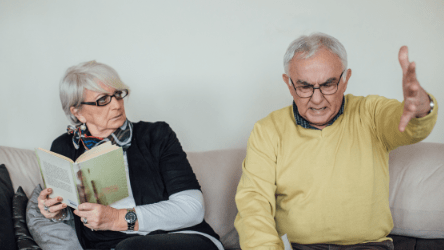 An elderly man sitting next to an elderly woman on a sofa. The elderly man looks mad.