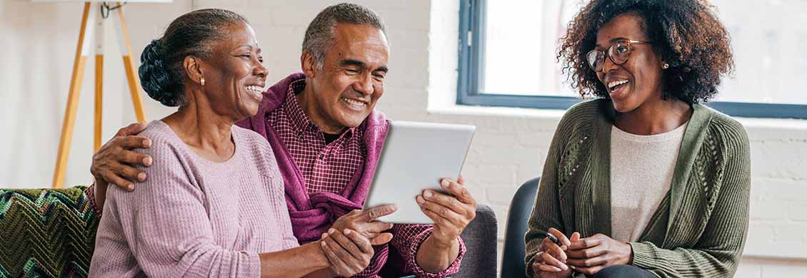 Mid-age female caregiver discusses home care agencies, like Visiting Angels, with a senior couple, holding a computer tablet.