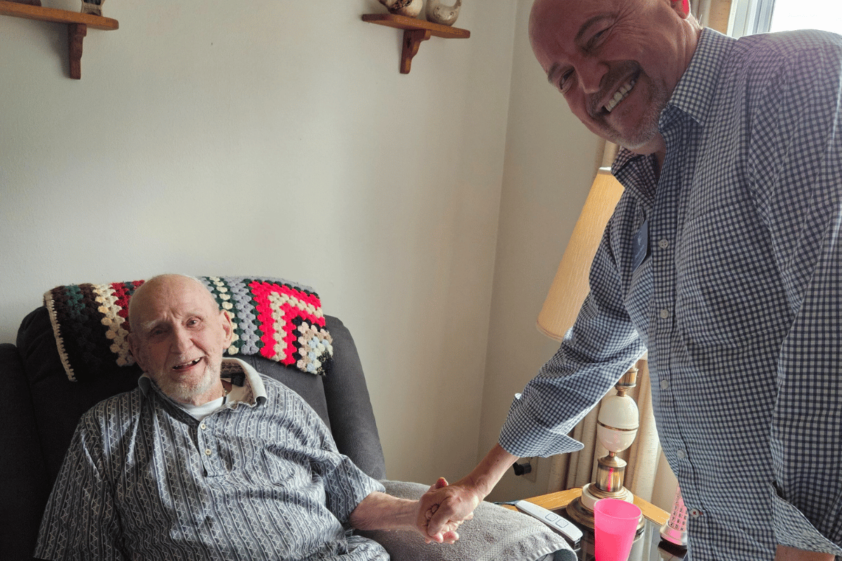 Denny Stultz grasps the hand of Visiting Angels of Southwest Michigan, Tony, who is sitting in a recliner, both are smiling at the camera. 