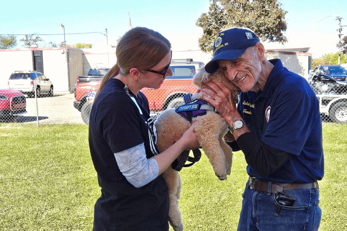 A Kalamazoo Veteran in a blue shirt and hat pets and nuzzles a Visiting Angels Therapy dog held by a volunteer in a blue shirt with long hair.