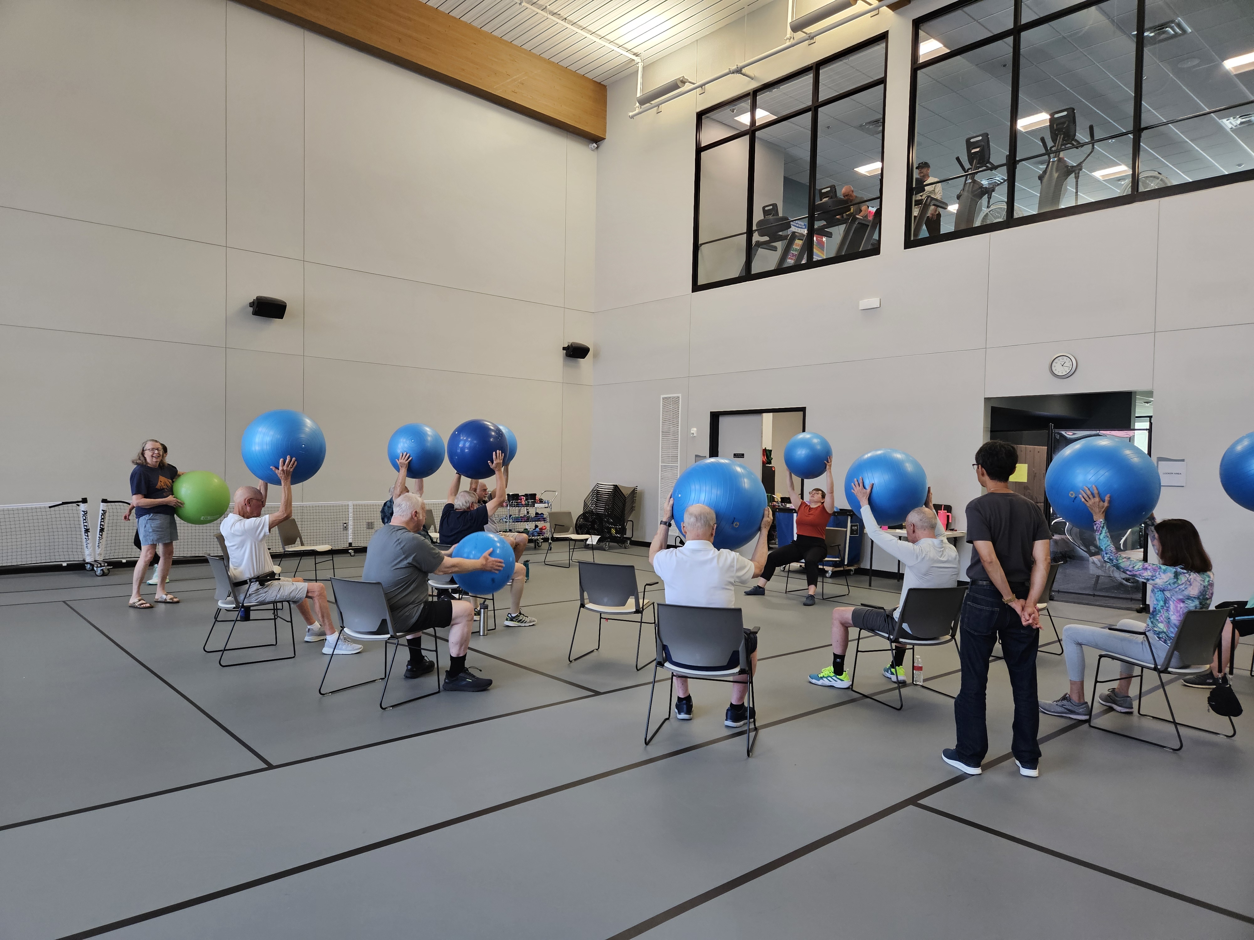 A group of individuals sit in chairs facing away from the camera and lift blue exercise balls over their heads as instructed in their Parkinson's Exercise class in a large room of the Portage Zhang Senior Center. 