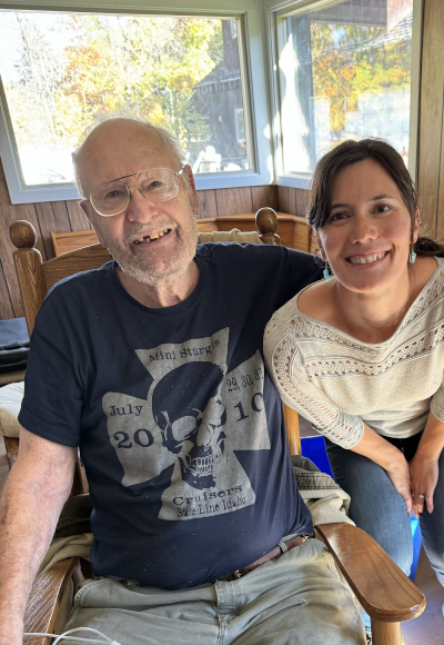 A client at Visiting Angels of Spokane, wearing a blue shirt and glasses, sitting comfortably in a chair with his arm around the owner, who is wearing a white shirt. Both individuals are smiling.