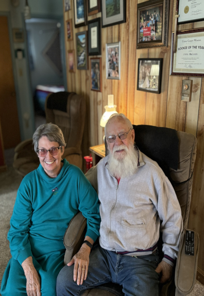Client of Visiting Angels of Spokane, dressed in a grey shirt, sits comfortably in a chair with his arm around his wife, who is wearing a blue shirt. Both individuals are smiling.