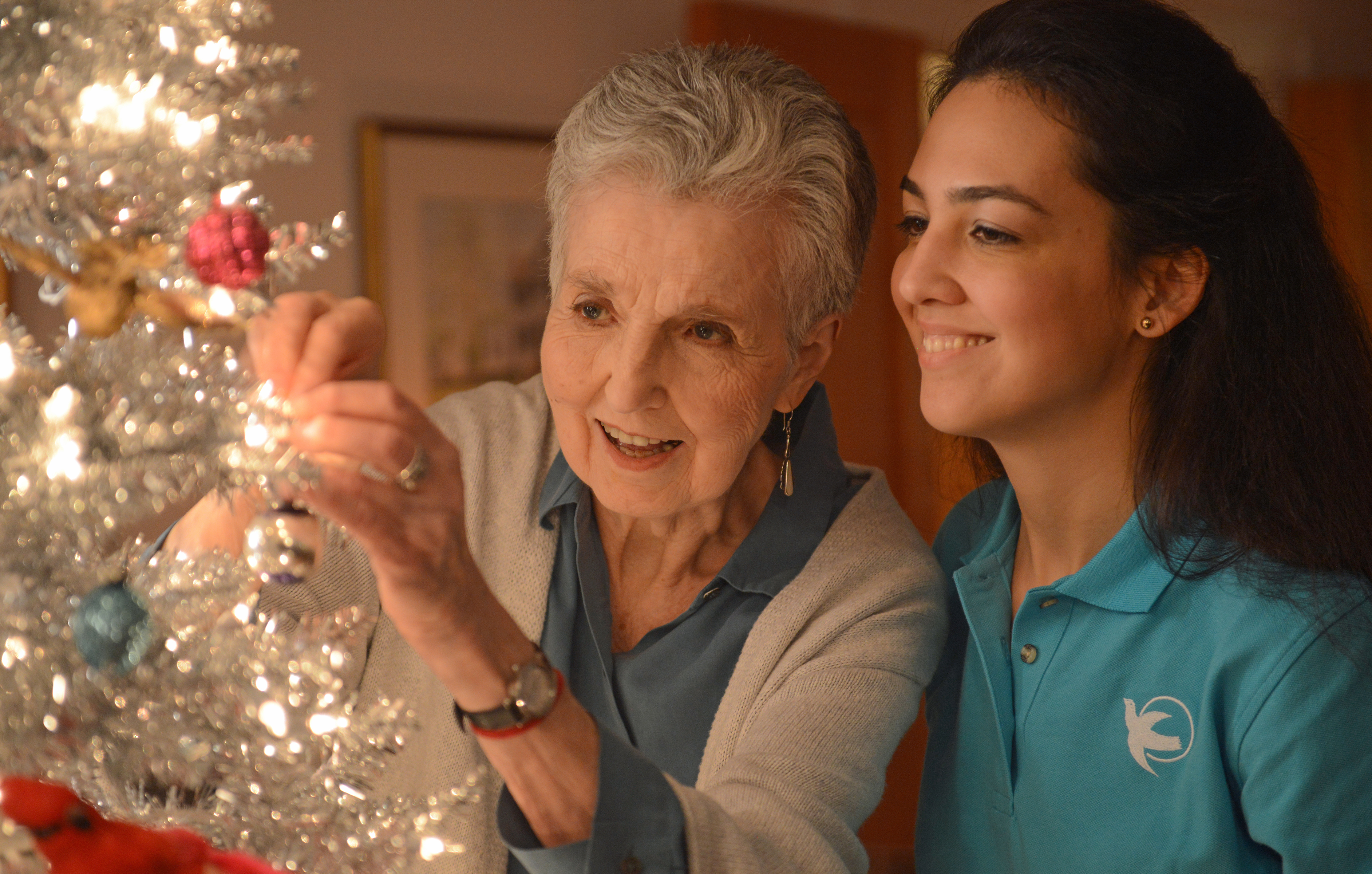 An elderly woman and their caregiver who has long brown hair and a teal Visiting Angels shirt hang ornaments on a silver christmas tree